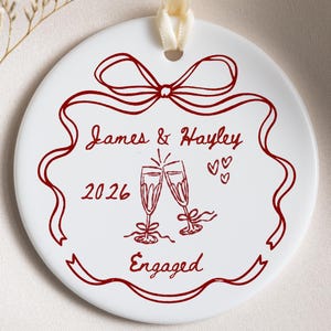May include: A round white ceramic ornament featuring a red line drawing of a ribbon bow, two champagne flutes, and three small hearts. The text reads James & Hayley, 2026, and Engaged. A cream-colored ribbon is attached to the top for hanging.