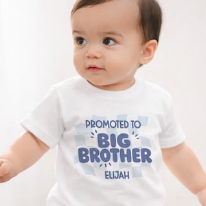Könnte beinhalten: Ein weißes T-Shirt mit dem Text "Promoted to Big Brother Elijah" in blauen Buchstaben. Der Text ist vor einem hellblauen Karomuster platziert. Das T-Shirt wird von einem Baby getragen.