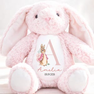 May include: A soft, pink plush bunny with long ears and white paws. The bunny has a white patch on its belly with a letter "A", a watercolor illustration of a bunny, the name "Amelia", and the date "08.04.2026".