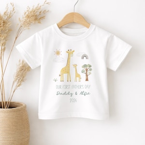 May include: White t-shirt with a giraffe family illustration, sun, rainbow, and tree. Text reads "OUR FIRST FATHER'S DAY Daddy & Alfie 2026". The shirt is hanging on a wooden hanger.