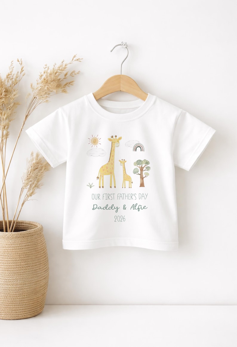 May include: White t-shirt with a giraffe family illustration, sun, rainbow, and tree. Text reads "OUR FIRST FATHER'S DAY Daddy & Alfie 2026". The shirt is hanging on a wooden hanger.