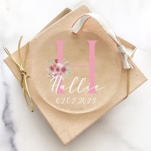 Op de afbeelding: Een transparante glazen ornament met een roze letter H, een bloemenontwerp en de naam Hallie met de datum 02.02.2023. De ornament is in een bruine doos met een gouden lint.