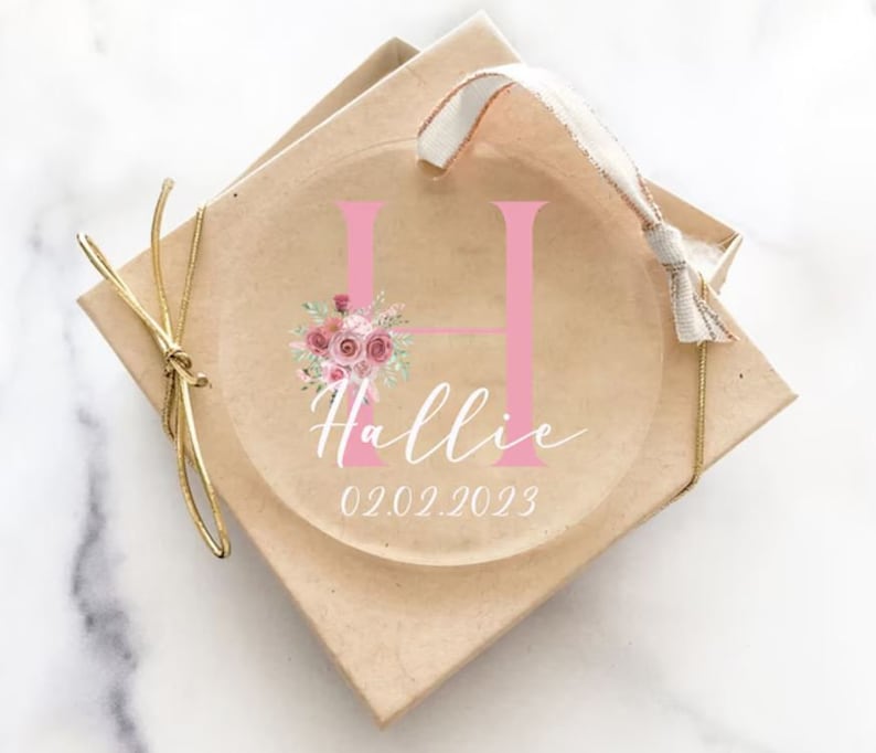 Op de afbeelding: Een transparante glazen ornament met een roze letter H, een bloemenontwerp en de naam Hallie met de datum 02.02.2023. De ornament is in een bruine doos met een gouden lint.