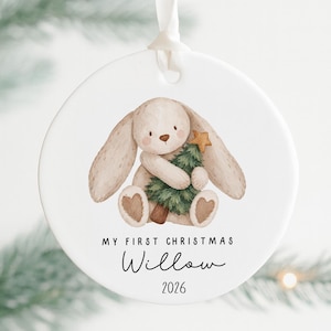 May include: A round white ceramic ornament featuring a watercolor illustration of a beige bunny holding a green Christmas tree with a gold star. The text reads MY FIRST CHRISTMAS, the name Willow, and the year 2026. A white ribbon is attached.