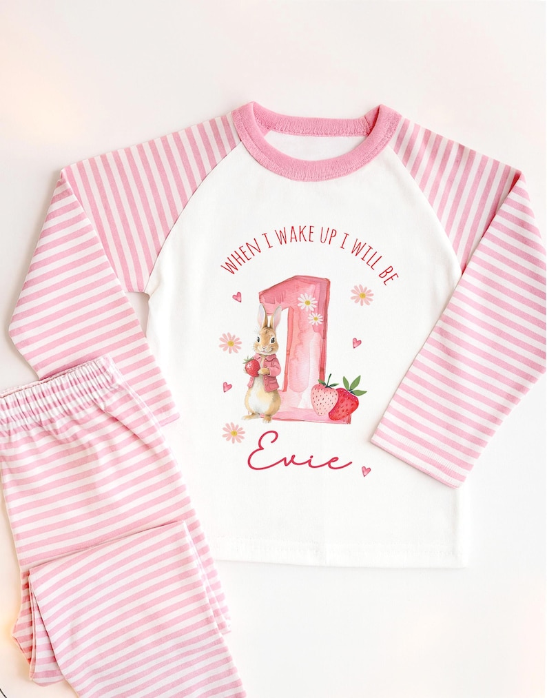 May include: A two-piece pajama set with a white top and pink striped sleeves and pants. The top features a pink number 1 with a bunny illustration, strawberries, and the text "WHEN I WAKE UP I WILL BE Evie."
