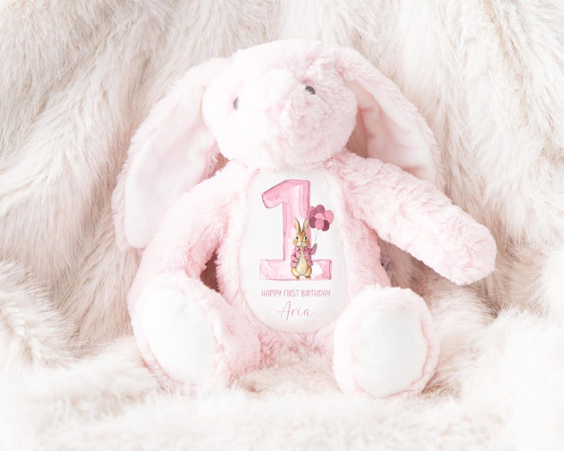 May include: Pink plush bunny rabbit with a white embroidered patch featuring a number one, a bunny rabbit illustration, and the text "Happy First Birthday Aria".