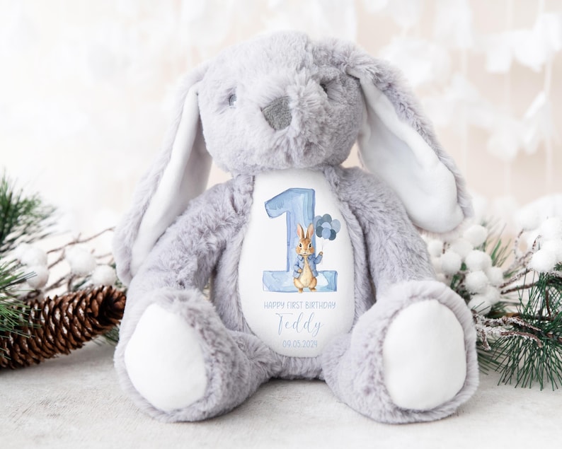 May include: A gray plush bunny with long ears, holding a white patch with a blue number one, a cartoon bunny, and the text "Happy First Birthday Teddy 09.05.2023".