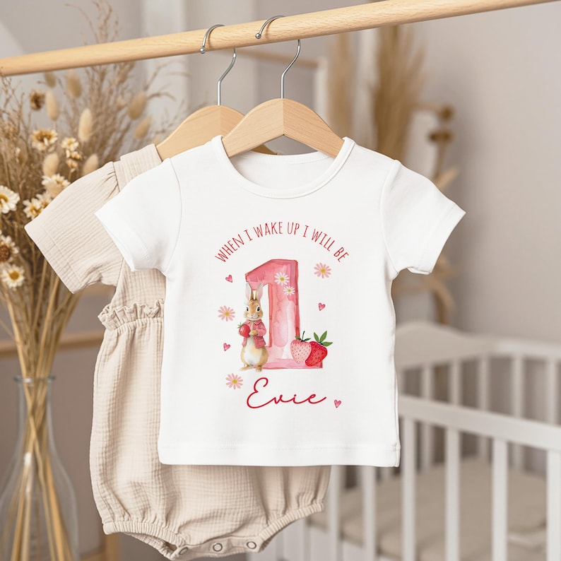 May include: White t-shirt with the text "WHEN I WAKE UP I WILL BE" in a circular pattern. The number "1" is pink with floral accents, and a bunny holding strawberries is featured. The name "Evie" is written below.