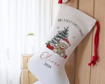 May include: White Christmas stocking with a red drawstring, featuring a cartoon bear wearing a Santa hat, a Christmas tree, and the text "My First Christmas Oliver 2024".