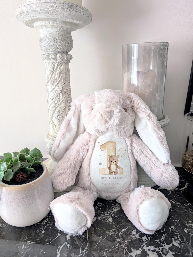 Op de afbeelding: Een roze pluche konijn met lange oren, met een patch met een nummer &eacute;&eacute;n, een beer en de tekst "Happy First Birthday Jensen 15.08.2021".