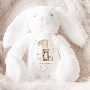 Op de afbeelding: Een witte pluche bunny met een bruine beer en het nummer &eacute;&eacute;n op zijn borst. De bunny heeft een gepersonaliseerd bericht met de tekst "Happy First Birthday Hudson 08.08.2024".