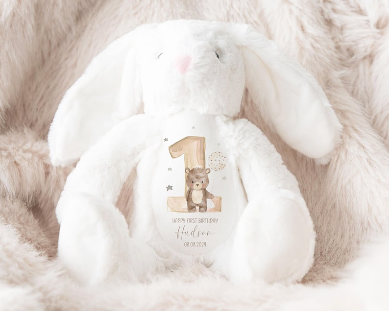 Op de afbeelding: Een witte pluche bunny met een bruine beer en het nummer &eacute;&eacute;n op zijn borst. De bunny heeft een gepersonaliseerd bericht met de tekst "Happy First Birthday Hudson 08.08.2024".