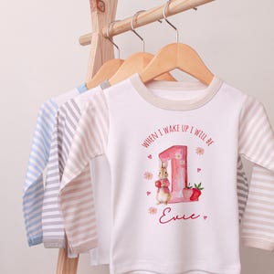 May include: White long-sleeved baby shirt with a bunny and strawberry graphic. The shirt has the text "WHEN I WAKE UP I WILL BE" and a name. Other shirts with striped sleeves hang on a wooden rack.