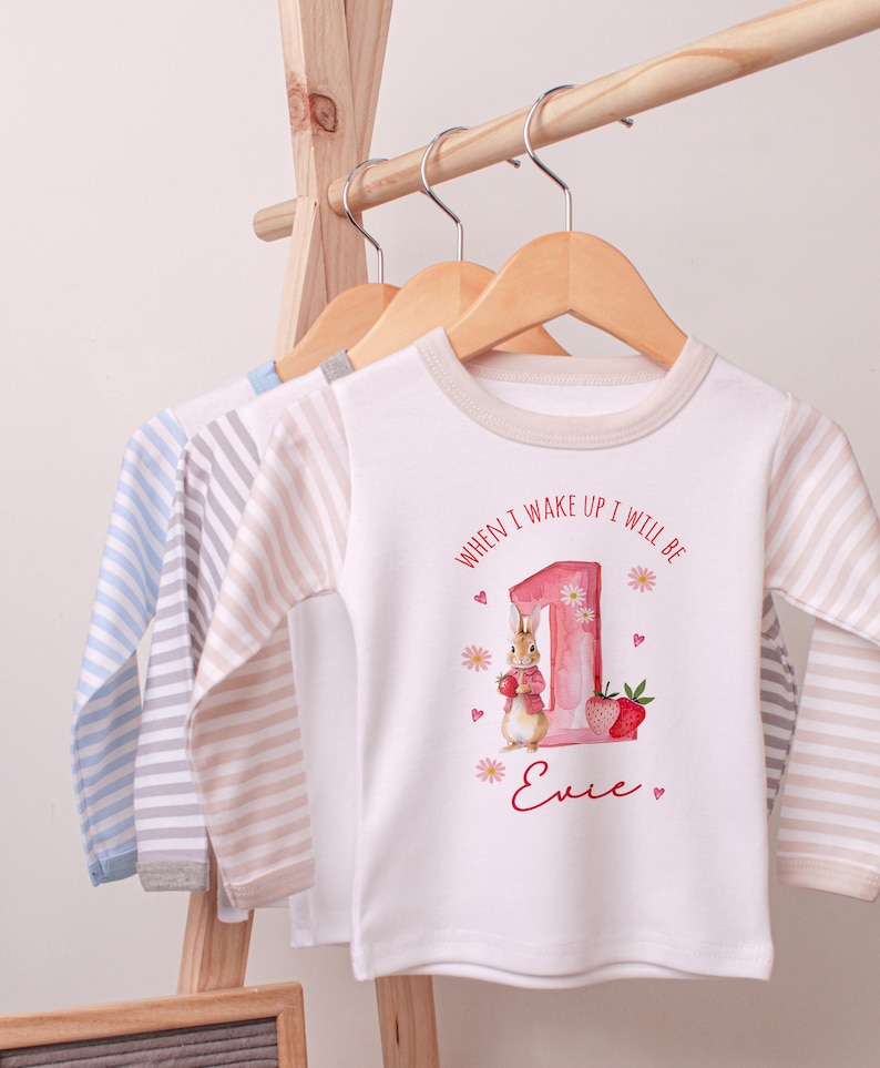 May include: White long-sleeved baby shirt with a bunny and strawberry graphic. The shirt has the text "WHEN I WAKE UP I WILL BE" and a name. Other shirts with striped sleeves hang on a wooden rack.