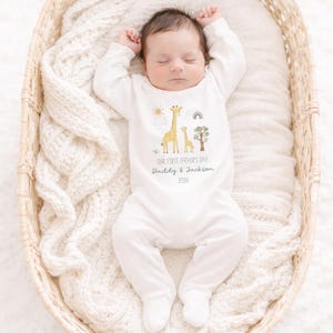 May include: A sleeping baby wearing a white onesie with a giraffe and tree graphic. The onesie has the text "OUR FIRST FATHER'S DAY Daddy & Jackson 2026". The baby is lying in a woven basket on a white blanket.