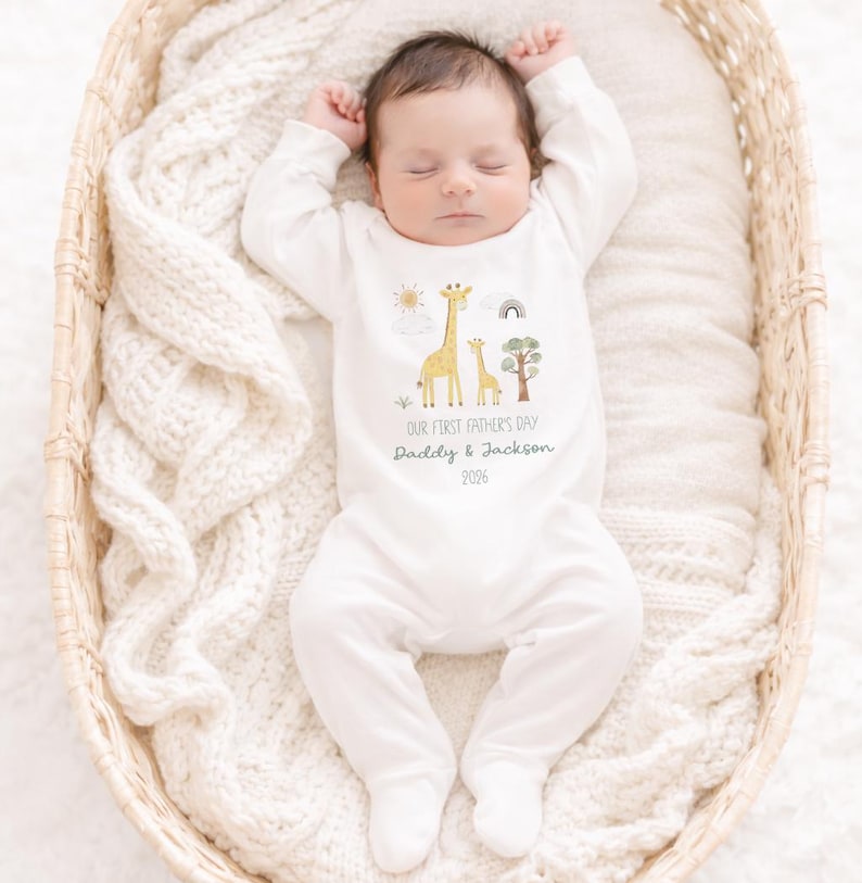 May include: A sleeping baby wearing a white onesie with a giraffe and tree graphic. The onesie has the text "OUR FIRST FATHER'S DAY Daddy & Jackson 2026". The baby is lying in a woven basket on a white blanket.