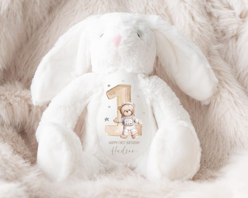 May include: White plush bunny with a brown number one and a bear astronaut design. The bunny has a personalized message that says "Happy First Birthday Hudson".