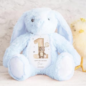 Op de afbeelding: Een lichtblauwe pluche bunny met lange oren, die een gepersonaliseerd patch vasthoudt met een bruine beer en het nummer &eacute;&eacute;n. Het patch luidt "Happy First Birthday Sam 02.06.2024".
