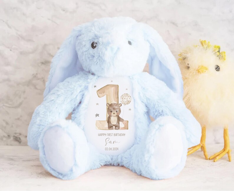 Op de afbeelding: Een lichtblauwe pluche bunny met lange oren, die een gepersonaliseerd patch vasthoudt met een bruine beer en het nummer &eacute;&eacute;n. Het patch luidt "Happy First Birthday Sam 02.06.2024".