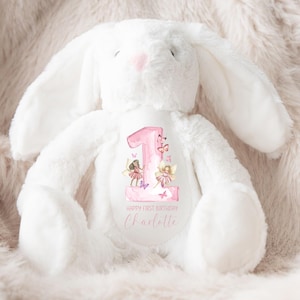 May include: White plush bunny with a pink number one and fairies on the front. The bunny has a pink nose and is sitting on a fluffy white surface. The text on the bunny says "Happy First Birthday Charlotte".
