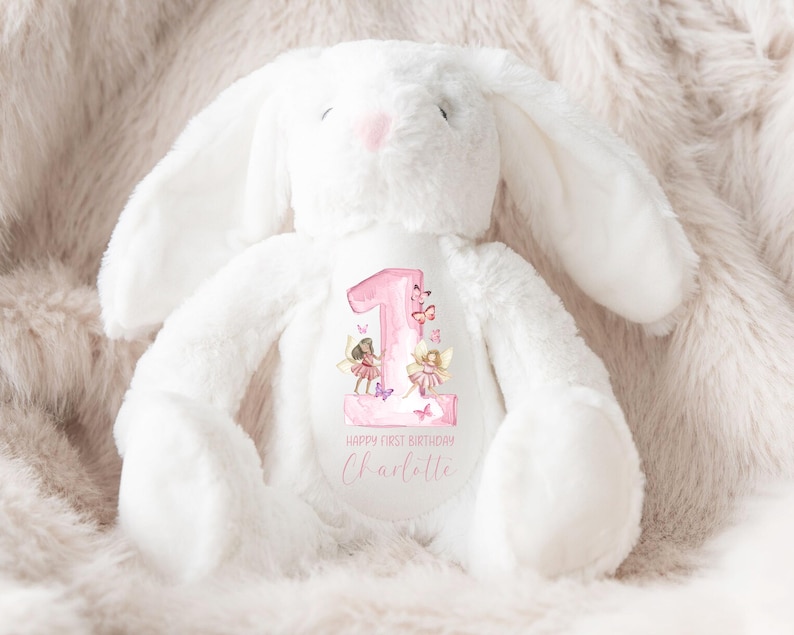 May include: White plush bunny with a pink number one and fairies on the front. The bunny has a pink nose and is sitting on a fluffy white surface. The text on the bunny says "Happy First Birthday Charlotte".