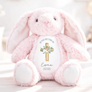 May include: A soft, pink plush bunny with long ears and white paws. The bunny has a cross design on its chest with the text "ON YOUR CHRISTENING DAY" above it, and the name "Esme" and date "01.03.2026" below.
