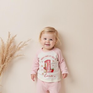 May include: A baby wearing a pink and white striped outfit. The long-sleeved top has a white front with a graphic of a bunny, the number 1, and the text "WHEN I WAKE UP I WILL BE" and "Evie". The outfit is for a first birthday.