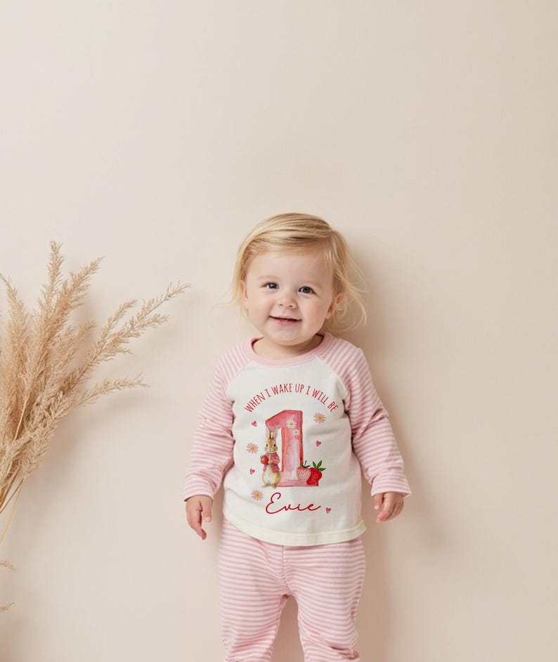 May include: A baby wearing a pink and white striped outfit. The long-sleeved top has a white front with a graphic of a bunny, the number 1, and the text "WHEN I WAKE UP I WILL BE" and "Evie". The outfit is for a first birthday.