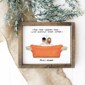 May include: A white framed print with an illustration of a couple sitting on an orange sofa in front of a fountain. The text "The One Where They Live Happily Ever After" is above the couple. The names "Rich & Amber" are below the couple.