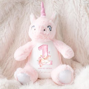 May include: Pink unicorn plush toy with a white embroidered patch that says "Happy First Birthday" and has a number "1" with a unicorn design. The unicorn is wearing a pink horn and has a white mane.