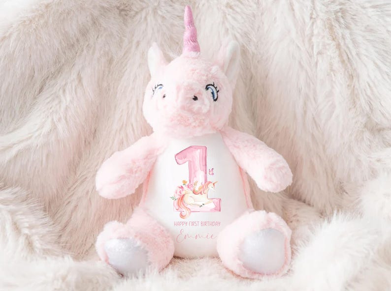 May include: Pink unicorn plush toy with a white embroidered patch that says "Happy First Birthday" and has a number "1" with a unicorn design. The unicorn is wearing a pink horn and has a white mane.