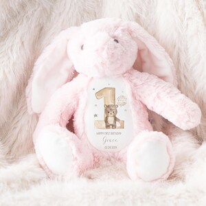 Op de afbeelding: Roze pluche konijn met een witte vlek op zijn borst. De vlek heeft een afbeelding van een bruine beer en de tekst "Happy First Birthday Grace 02.04.2024".