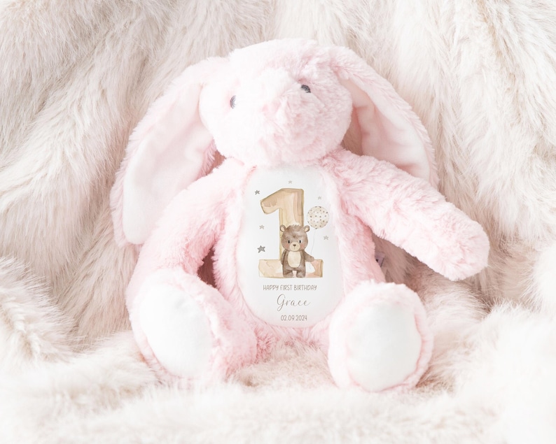 Op de afbeelding: Roze pluche konijn met een witte vlek op zijn borst. De vlek heeft een afbeelding van een bruine beer en de tekst "Happy First Birthday Grace 02.04.2024".