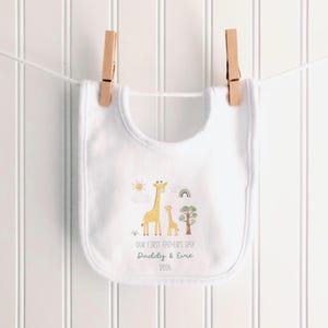 May include: White baby bib with a giraffe family illustration and the text "OUR FIRST FATHER'S DAY Daddy & Evie 2026". The bib is hanging from a clothesline with two wooden clothespins.
