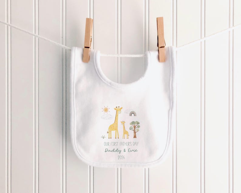 May include: White baby bib with a giraffe family illustration and the text "OUR FIRST FATHER'S DAY Daddy & Evie 2026". The bib is hanging from a clothesline with two wooden clothespins.
