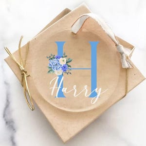 Op de afbeelding: Een transparante glazen kerstbal met een blauwe letter H en een bloemmotief. De naam Harry staat in witte cursieve letters onder de letter. De kerstbal zit in een bruine doos met een gouden lint.