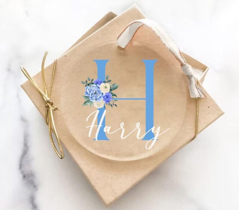 Op de afbeelding: Een transparante glazen kerstbal met een blauwe letter H en een bloemmotief. De naam Harry staat in witte cursieve letters onder de letter. De kerstbal zit in een bruine doos met een gouden lint.