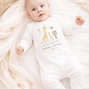 May include: White baby onesie with long sleeves and feet. The onesie features a giraffe family graphic with a sun, rainbow, and tree. Text reads "OUR FIRST FATHER'S DAY Daddy & Tommy 2026". The baby is lying on a white blanket.