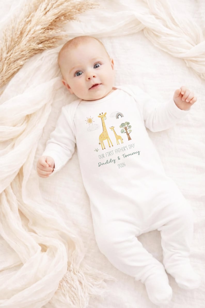 May include: White baby onesie with long sleeves and feet. The onesie features a giraffe family graphic with a sun, rainbow, and tree. Text reads "OUR FIRST FATHER'S DAY Daddy & Tommy 2026". The baby is lying on a white blanket.