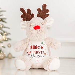 May include: A beige plush reindeer toy with brown antlers and a red nose. The reindeer is wearing a white shirt with the text "Albie's First Christmas 2024" in red and black.