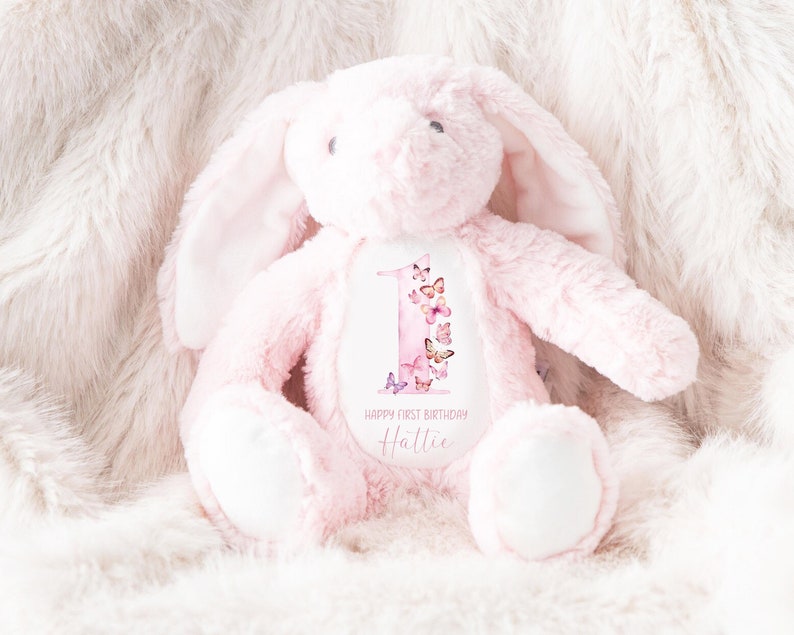 May include: Pink plush bunny with a white patch on its chest. The patch features a watercolor-style number one with butterflies and the text "Happy First Birthday Hattie".