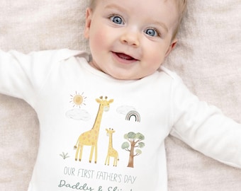 Op de afbeelding: Een witte baby-onesie met lange mouwen en een aquarelontwerp van giraffen, zon, wolken, regenboog en boom. De tekst "OUR FIRST FATHER'S DAY Daddy & Elijah 2026" staat eronder. De baby lacht.