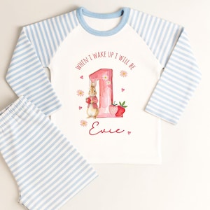 May include: White and blue striped pajama set with a long-sleeved top and shorts. The top features a graphic of a bunny, a number 1, and the text "WHEN I WAKE UP I WILL BE" and a name. The sleeves are blue and white striped.