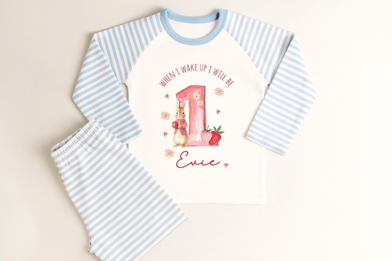 May include: White and blue striped pajama set with a long-sleeved top and shorts. The top features a graphic of a bunny, a number 1, and the text "WHEN I WAKE UP I WILL BE" and a name. The sleeves are blue and white striped.