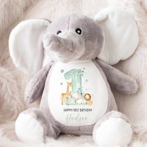 May include: A gray plush elephant stuffed animal with white ears and a white belly. The belly has a white patch with a green number one, a giraffe, an elephant, and a lion. The text "Happy First Birthday Hudson" is printed on the patch.
