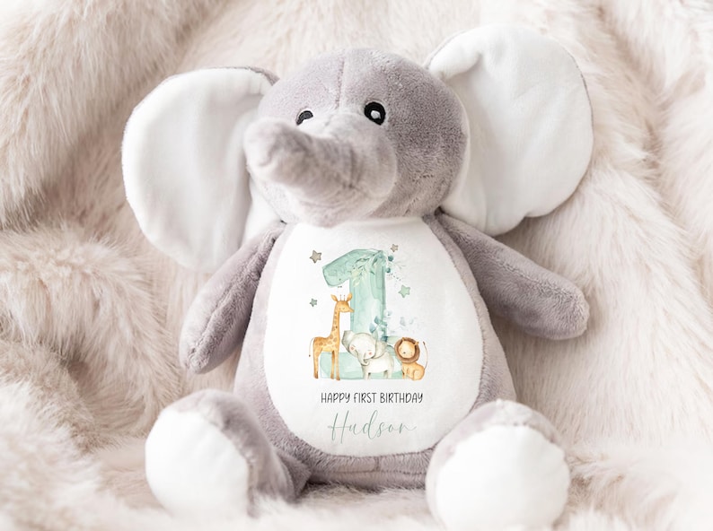 May include: A gray plush elephant stuffed animal with white ears and a white belly. The belly has a white patch with a green number one, a giraffe, an elephant, and a lion. The text "Happy First Birthday Hudson" is printed on the patch.