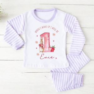 May include: White and lavender striped pajama set for a child. The top features a graphic of a bunny, the number 1, and the text "When I wake up I will be 1". The name "Evie" is written in red script below the graphic.