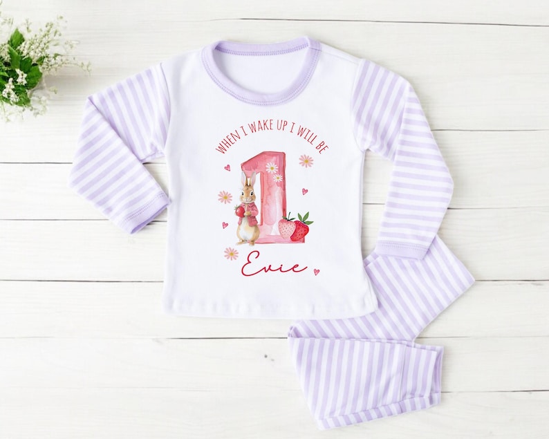 May include: White and lavender striped pajama set for a child. The top features a graphic of a bunny, the number 1, and the text "When I wake up I will be 1". The name "Evie" is written in red script below the graphic.