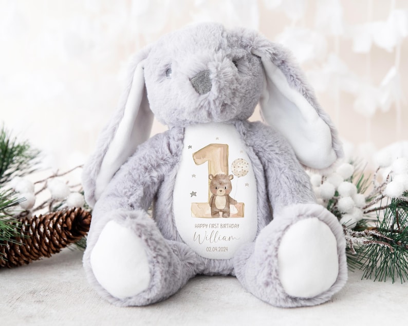 Op de afbeelding: Grijze pluche konijn met een witte vlek op zijn borst. De vlek heeft een bruine beer die een ballon vasthoudt en een houten nummer &eacute;&eacute;n met gouden sterren. De tekst "Happy First Birthday William 02.04.2024" staat op de vlek.