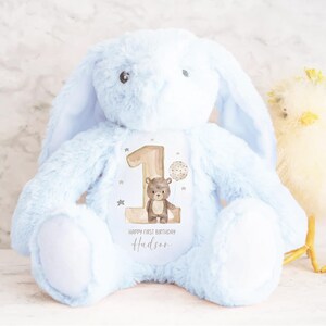 May include: A light blue plush bunny with a personalized patch that says "Happy First Birthday Hudson" and features a brown bear with a balloon. The bunny is sitting on a white surface.
